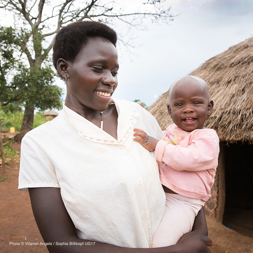 a mother holds and smiles at her baby in front of a hut in a Ugandan village; copyright Vitamin Angels / Sophia Billikopf UG17